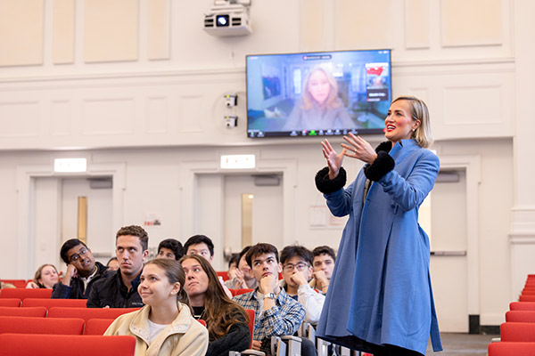 A woman in a lecture hall with students seated looks up at a screen showing another women.