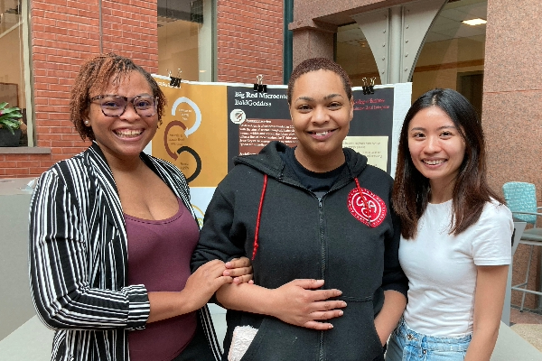 Molebi, JewleKrystal and Sheryl stand in front of a BaldGoddess trifold in Sage Hall's atrium.