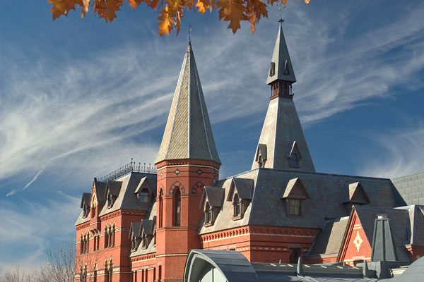 Facade of Sage Hall with autumn leaves visible in the foreground and a blue sky with wispy clouds in the background.