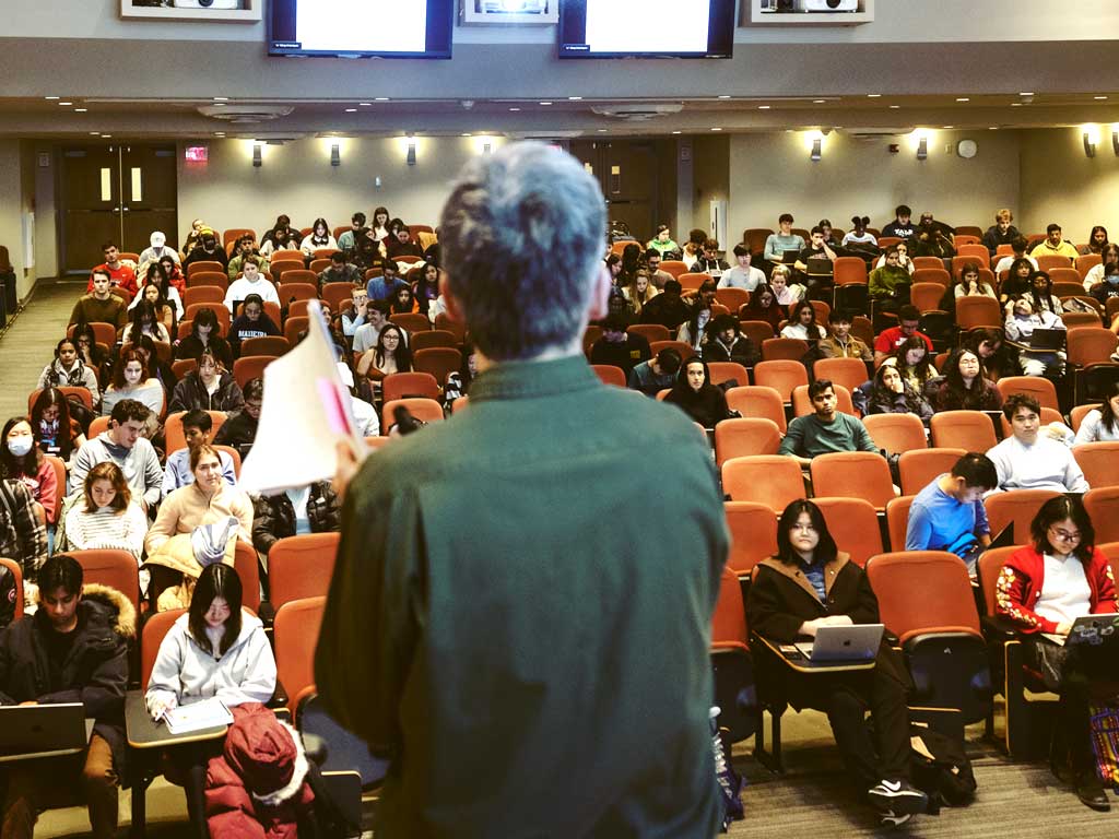 A person in a green shirt stands in front of a classroom giving a lecture.