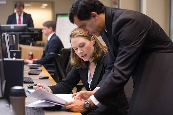 A woman in business attire sits and looks at papers with a man standing next to her.