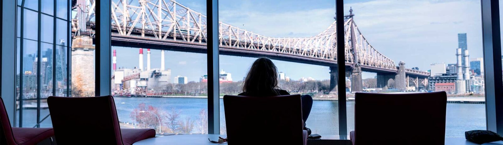 A person sits in a chair facing a window where a river and bridge are visible.