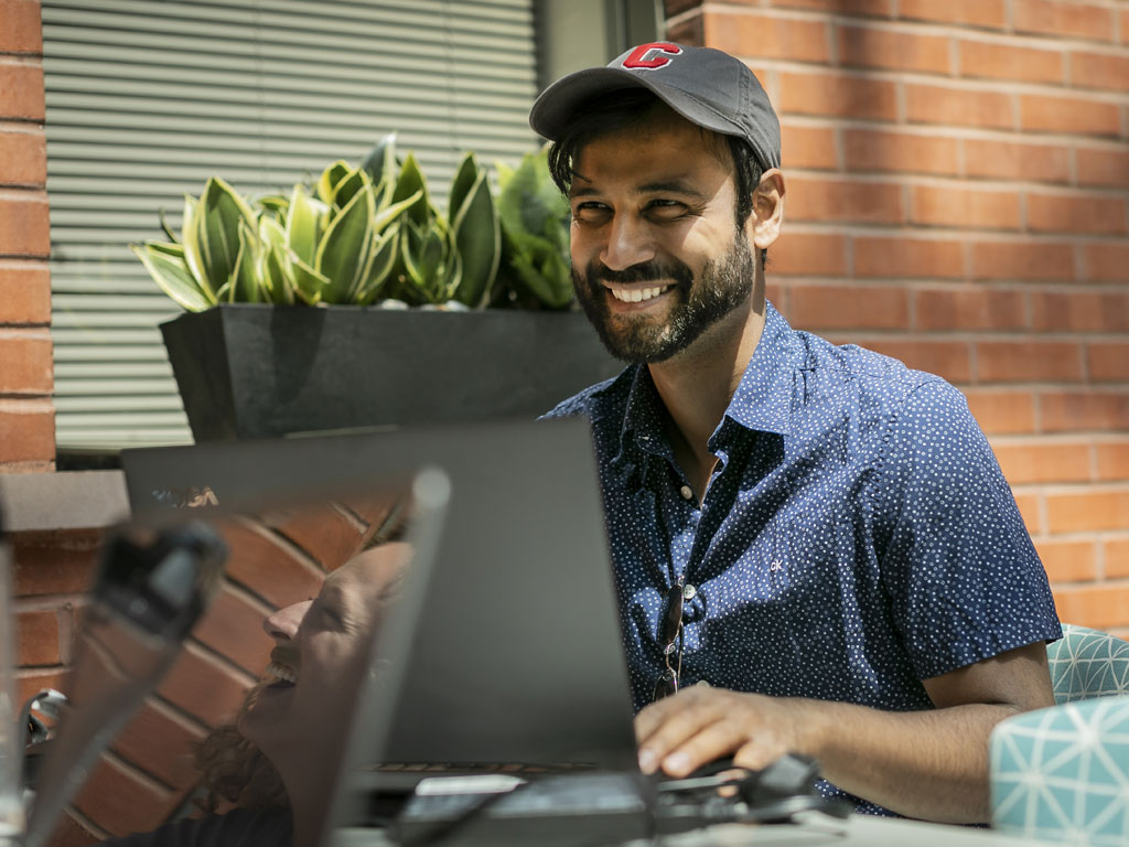 man wearing a baseball hat smiles with a laptop open in front of him.