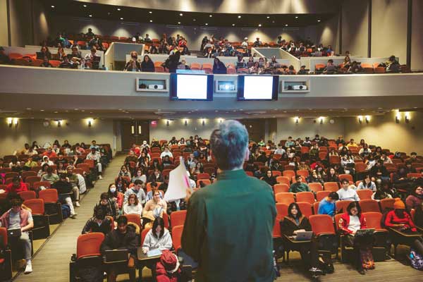 A professor stands in front of a large theater-style classroom.