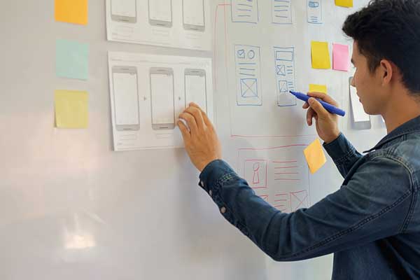 A person stands at a whiteboard drawing layouts for elements on an outline of a cell phone.