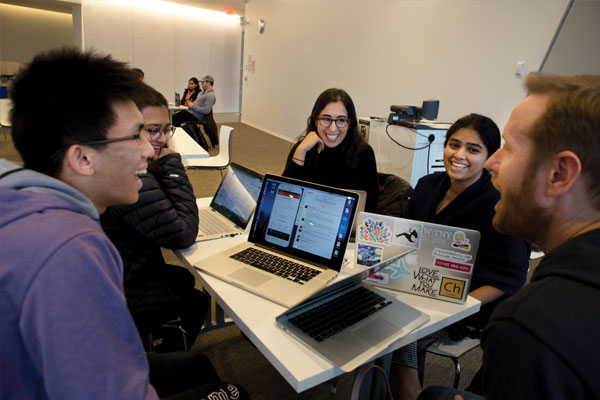 Students smile and laugh around a table with laptops open in front of them.