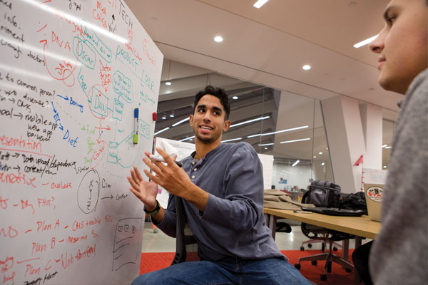 A student talks with his hands and gestures towards a whiteboard filled with writing.