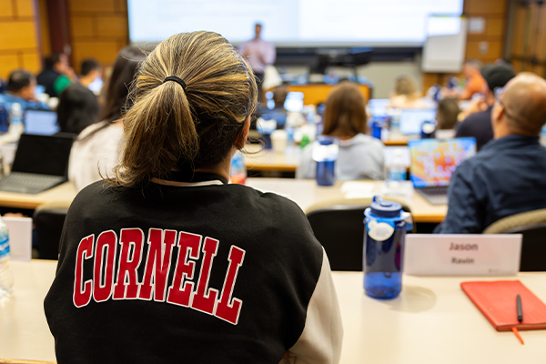 A student wearing a letterman jacket that says “Cornell” with a professor at the front of a room in the background.