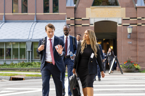 Students in business attire walk across a street with the facade of Sage Hall in the background.