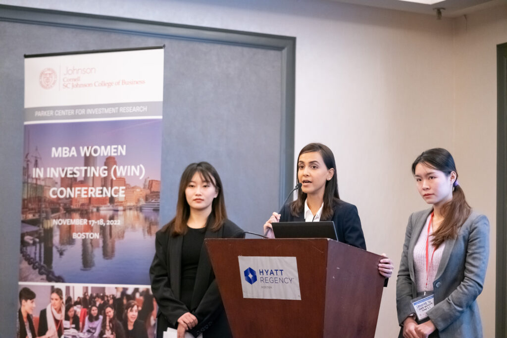 Three students presenting at the MBA Women in Investing Conference. One student is standing at the microphone podium, while the other two stand behind her.