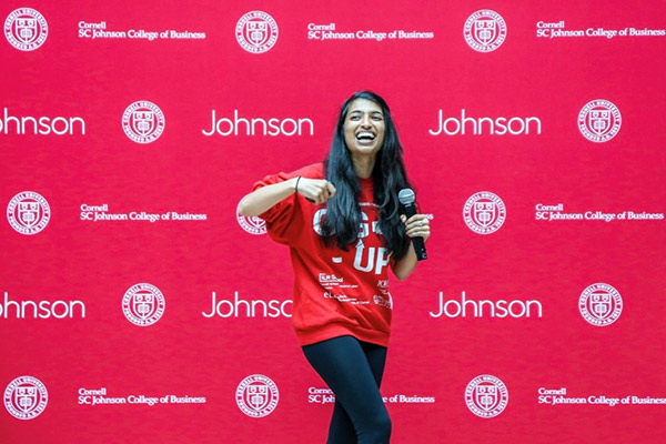 Trishala Dessai gesturing and smiling on stage wearing a red Gig-Up sweatshirt with a Johnson School banner in the background.