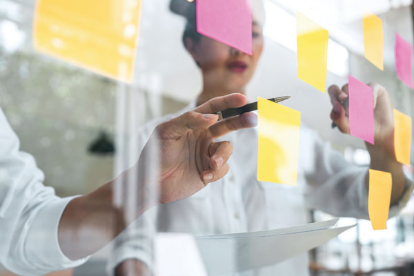 Two people writing on Post-it notes on a clear board.