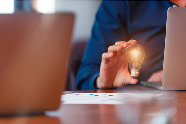 a hand resting on a table next to a laptop holding a brightly lit lightbulb.