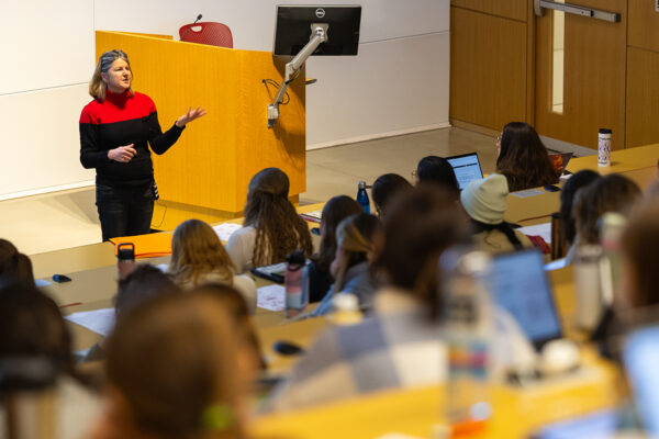 A woman professor lecturing in an auditorium style class room filled with students.