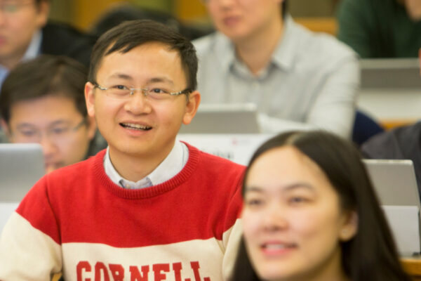 A student with a Cornell sweater on listening to a lecture with other students in the background.