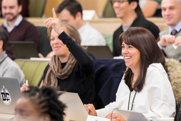 Two female students sitting in class. One raises her hand and the other smiles.