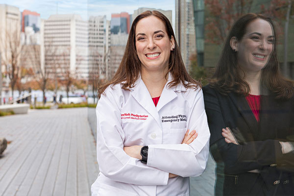 Image of a female health care professional in a white lab coat leaning against an exterior building wall in the city