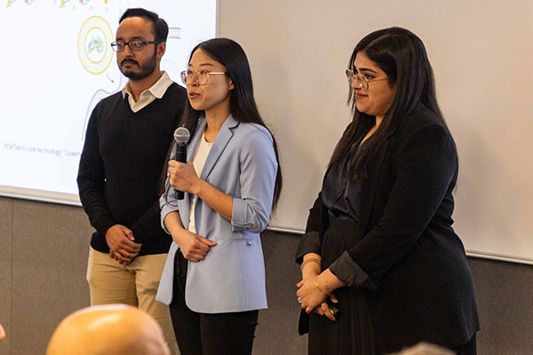 Two women and a man standing in th front of a room with a screen behind them. One woman is speaking into a microphone.