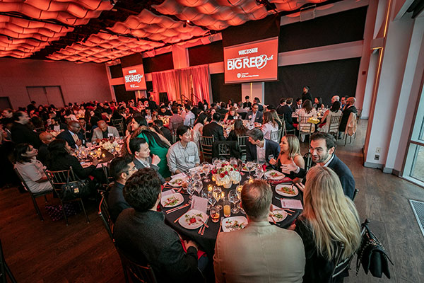 a large banquet hall filled with people seated at round tables and with two large red banners with the text Big Red Bash hanging on either side of the stage.