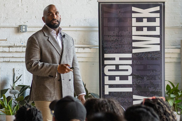 A man standing in front of a room full of people next to a Tech Week banner and speaking.