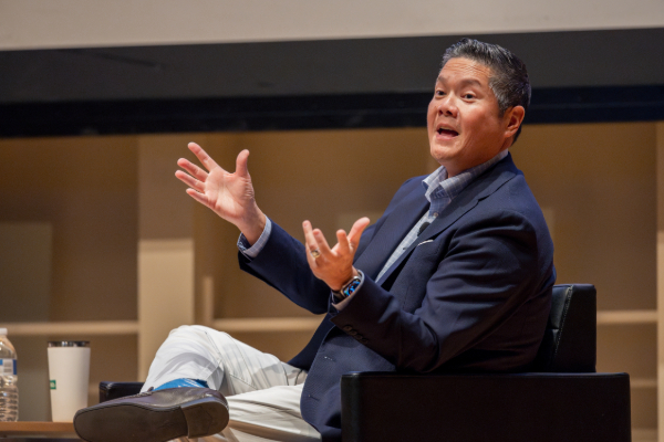 A man sits on stage talking with his hands outstretched.