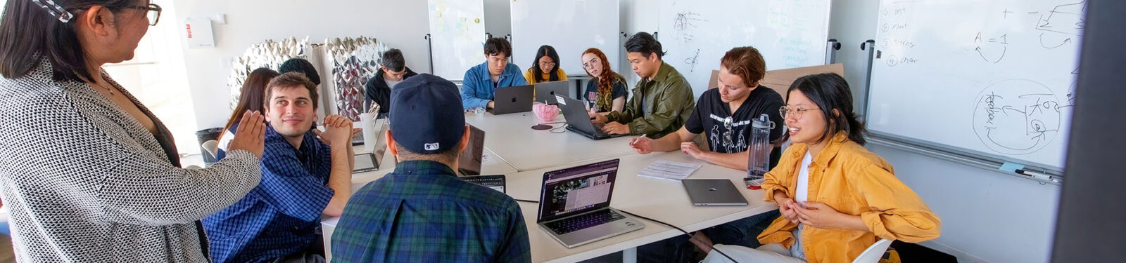 students with laptops listening to a professor speaking in a white room at a large table