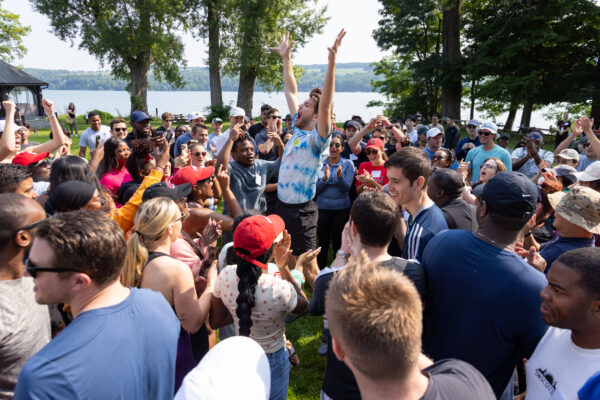Graduate business students dressed in casual active attire, gathered in a large crowd in a green space next to a lake and row of trees, looking towards the center where a young man in a tie-dyed t-shirt and headband is jumping very high while shouting