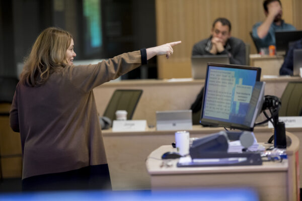 A professor stands at a computer in front of a classroom, with a partial view of the student audience.